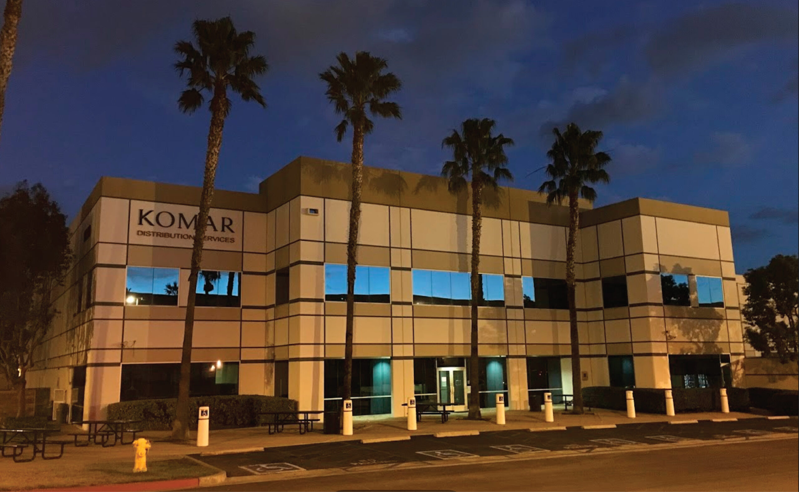 Komar Distribution office building at dusk with palm trees and illuminated windows.